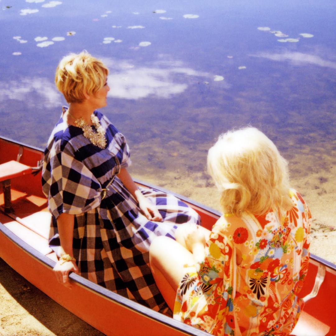 Two women sitting in a canoe on a calm lake with trees reflected on the water's surface, enjoying a peaceful day outdoors.