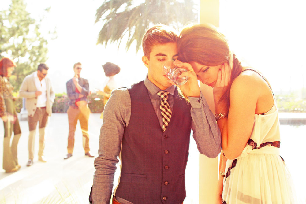 A stylish man and woman pose in bright sunlight, outdoors near a blurred gathering and pool; the man drinks from a glass.