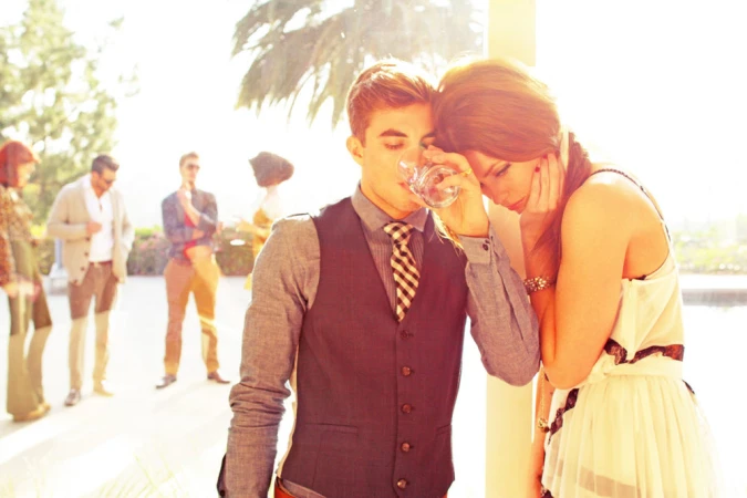 A stylish man and woman pose in bright sunlight, outdoors near a blurred gathering and pool; the man drinks from a glass.