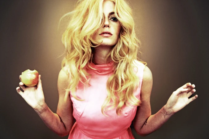 Blonde woman wearing a pink sleeveless dress holding a bitten apple against a dark gradient background