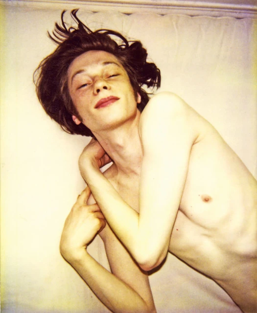 Artistic portrait of a bare-chested fair-skinned young adult lying on a white surface with a serene expression and eyes closed.