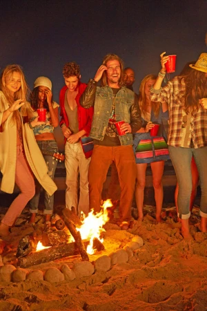 Group of friends gather around a bonfire on a sandy beach at night, holding red cups and wearing casual attire, creating a lively atmosphere.