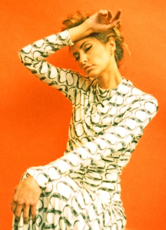 Woman in patterned long-sleeve dress with hand on forehead against a solid orange background