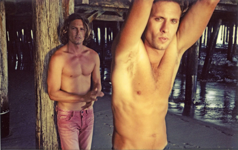 Two shirtless adult males pose on a beach beneath a wooden pier, one with arms raised, creating a sun-drenched, casual lifestyle image.