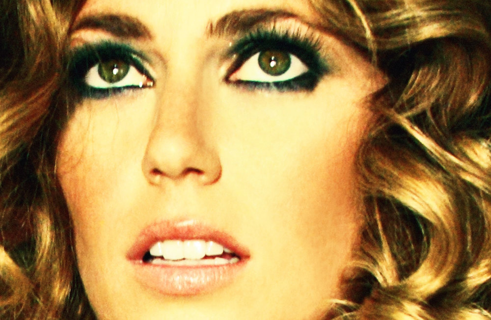 Close-up portrait of a young adult woman with curly blonde hair and green eyes wearing green eyeshadow and lipstick looking upwards.