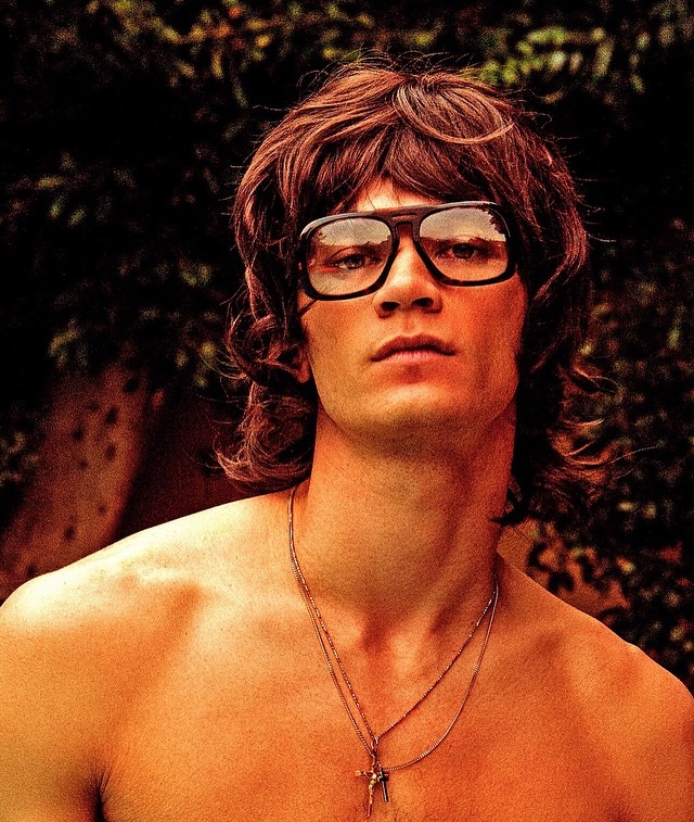 Portrait of a fair-skinned adult male with long brown hair wearing eyeglasses and a necklace against a leafy green backdrop.