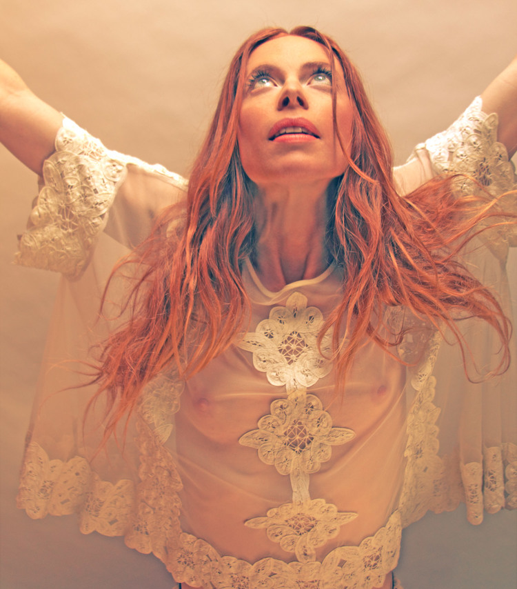 A woman with red hair in a lace-trimmed top gazes upwards with arms raised against a warm, muted background in a studio shot.