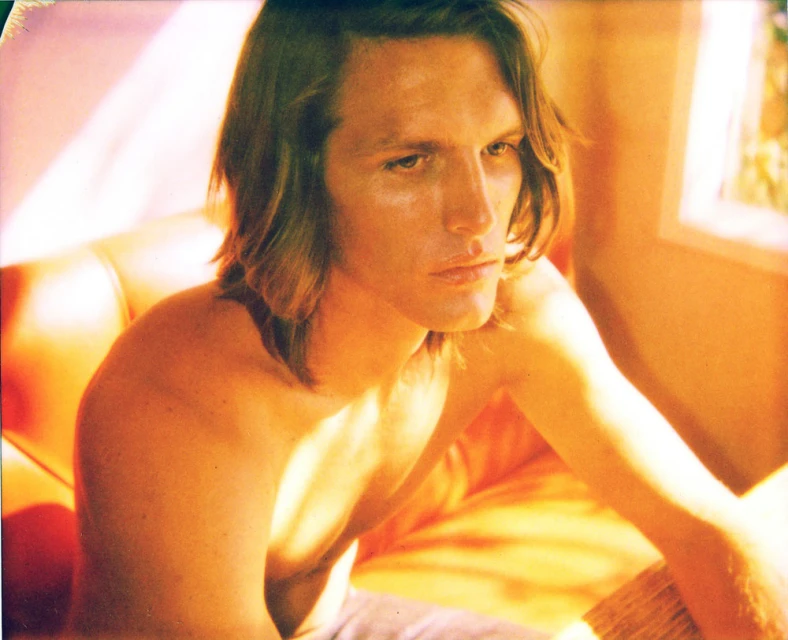 Portrait of an adult male with long brown hair, bare chest, and blue jeans sitting indoors near a window with natural lighting.