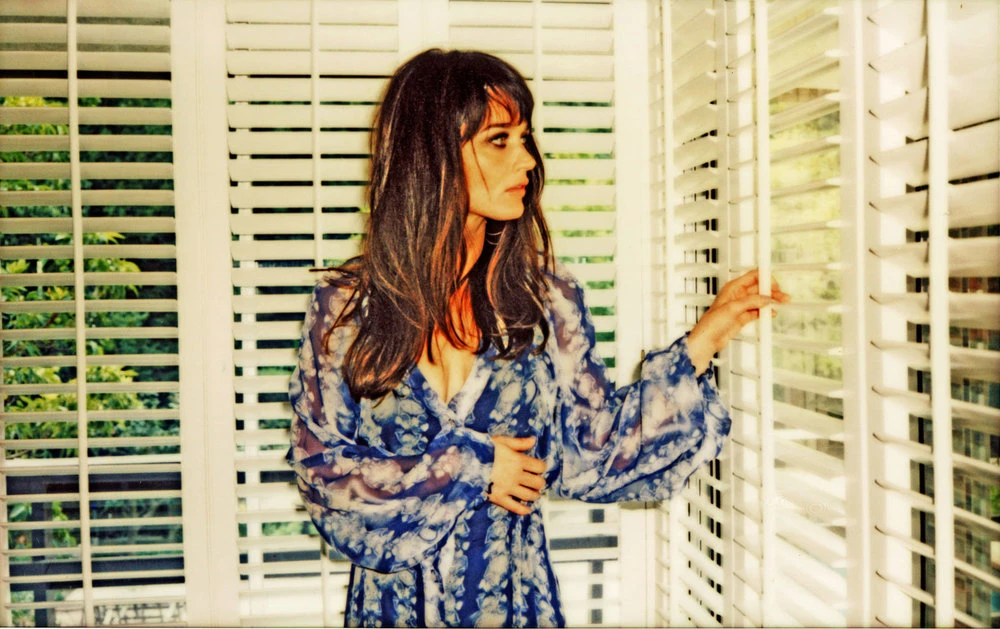 Attractive brunette woman in a blue and white patterned dress is standing indoors, looking out a window with white horizontal blinds.