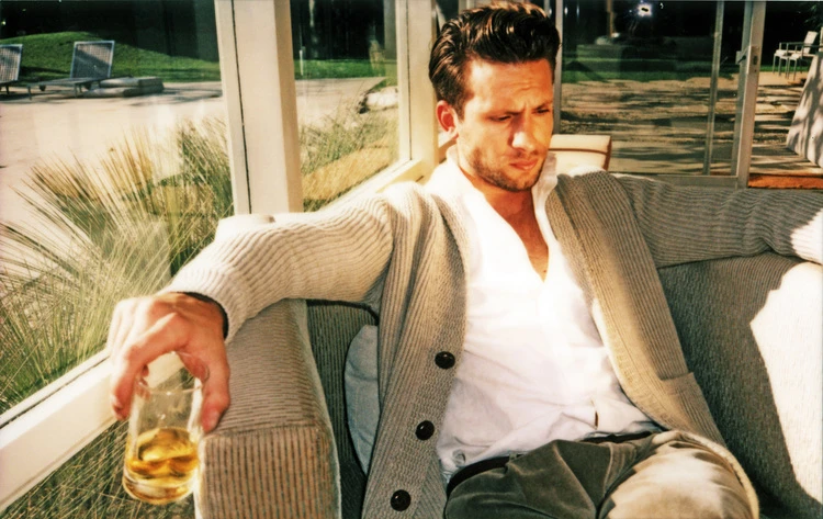 Indoor shot of a seated adult male holding a drink, wearing a cardigan sweater and collared shirt, in a sunlit area near a window.