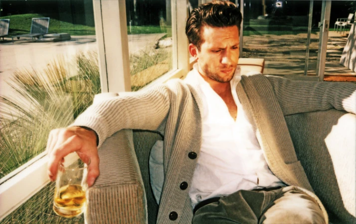 Indoor shot of a seated adult male holding a drink, wearing a cardigan sweater and collared shirt, in a sunlit area near a window.