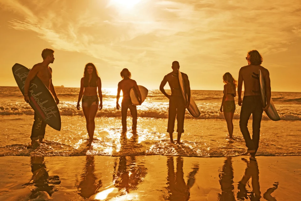 A group of six people, including men and women, stand on a beach at sunset holding surfboards, creating a warm, sunny coastal scene.