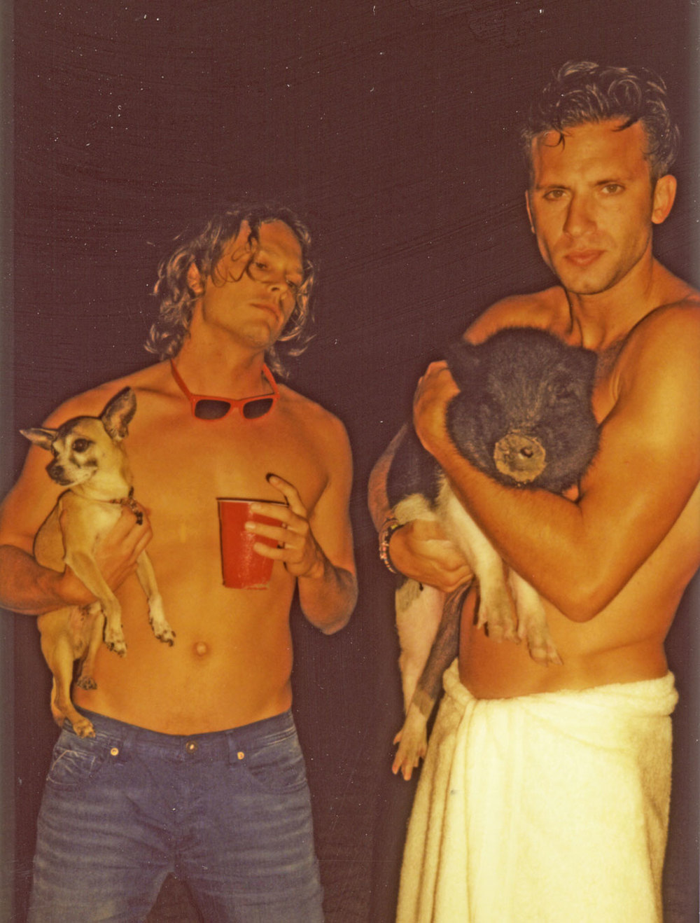 Two shirtless men pose outdoors, one holds a chihuahua and the other a pig, creating a candid portrait.