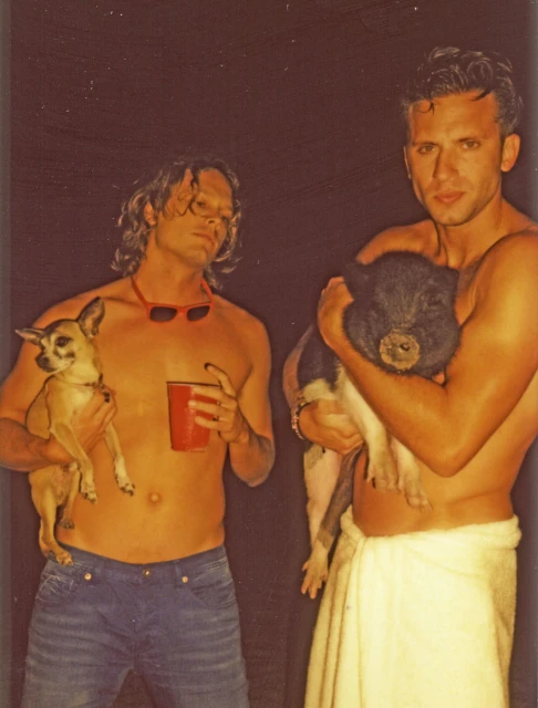 Two shirtless men pose outdoors, one holds a chihuahua and the other a pig, creating a candid portrait.