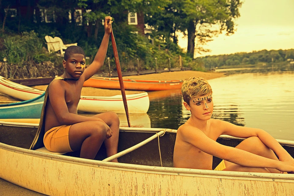 Two children with decorative face paint sit in a canoe on a lake with a beach and trees visible on the shoreline in the background.