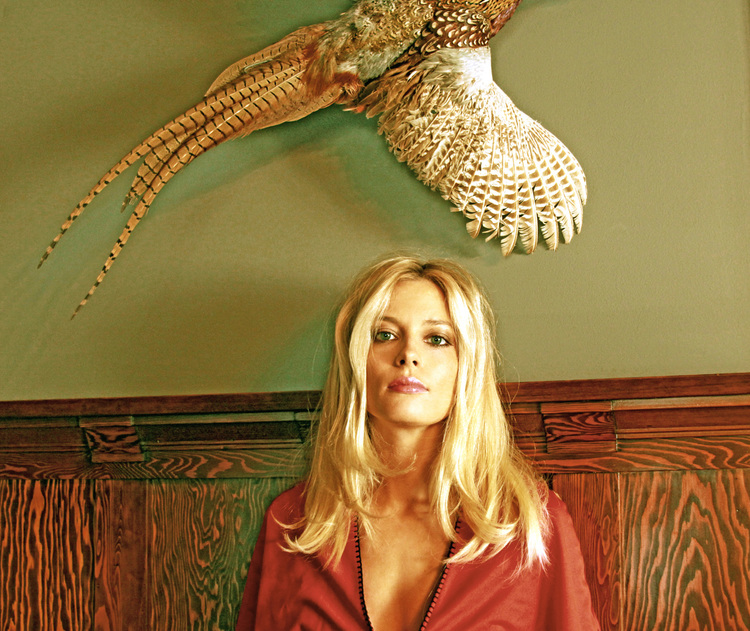 Portrait of a blonde woman in a red garment indoors with a mounted pheasant taxidermy on the wall as a vintage-style backdrop.