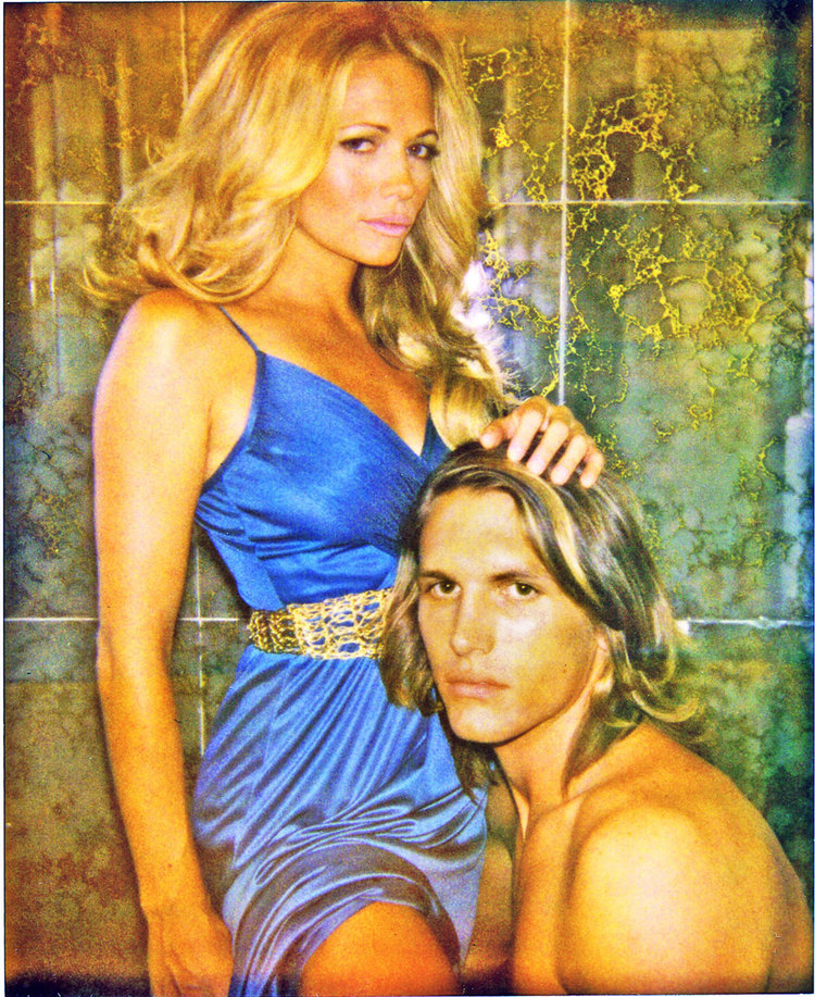 Blonde woman in blue dress with gold belt standing beside a shirtless man with long hair by Stephanie Vovas