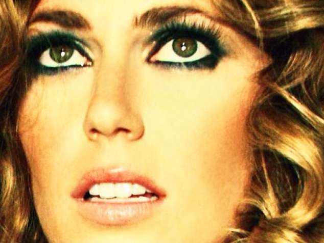 Close-up portrait of an adult woman with green eyes, dark green eye shadow, wavy blonde hair, and fair skin tone. Beauty shot.