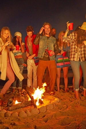 Group of young adults gather around a bonfire at night on a sandy beach, holding red cups, and enjoying an evening get-together.