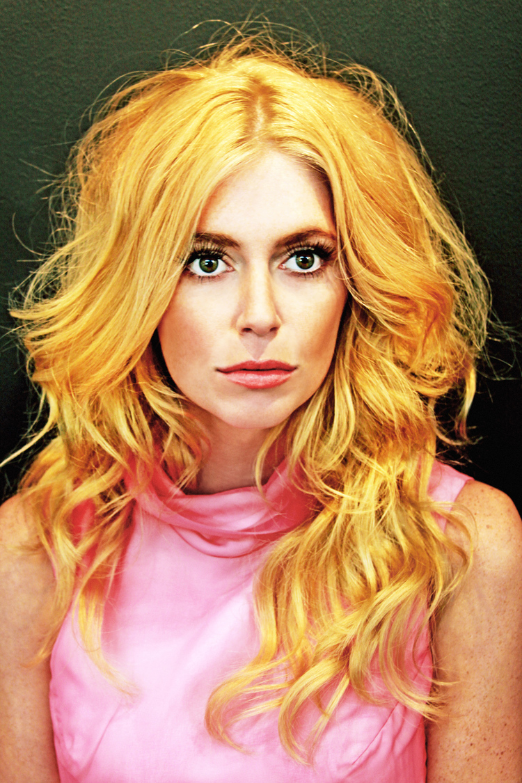 Close-up portrait of a woman with curly blonde hair and a pink dress against a dark background, showcasing a studio fashion style.