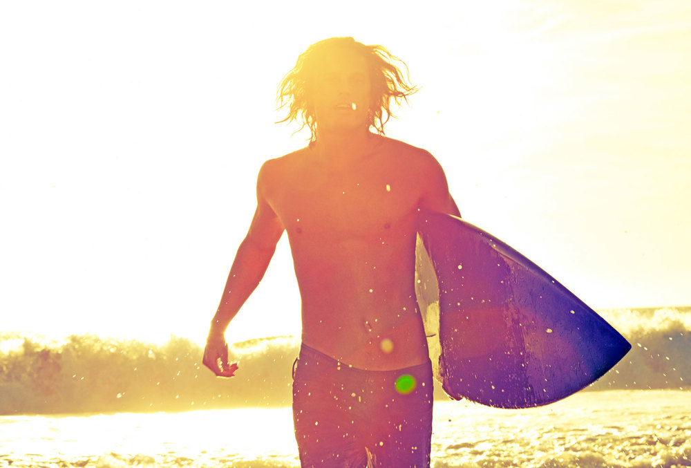 A shirtless man with long hair carries a surfboard on a sunny beach, illuminated by bright sunlight and waves in the background.