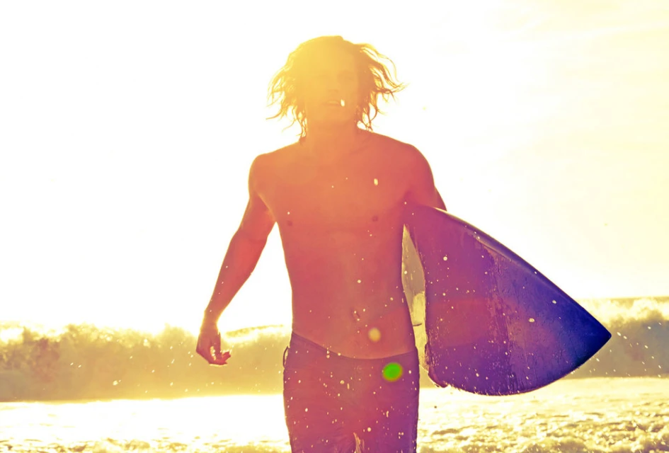 A shirtless man with long hair carries a surfboard on a sunny beach, illuminated by bright sunlight and waves in the background.
