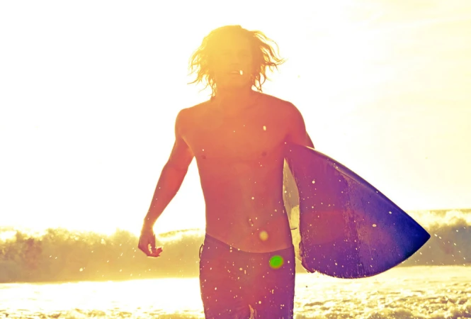 A shirtless man with long hair carries a surfboard on a sunny beach, illuminated by bright sunlight and waves in the background.