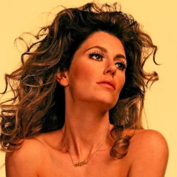 Close-up portrait of a woman with voluminous brown hair wearing a pendant necklace against a warm yellow background.