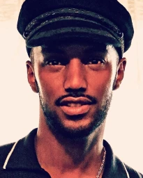 Close-up portrait of a man wearing a black captain hat and a dark polo shirt against a bright background.