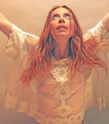 Red-haired woman in a sheer white lace blouse with arms raised, photographed by Stephanie Vovas against a neutral studio backdrop.