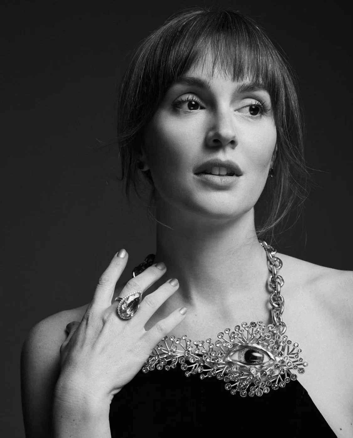 Portrait of a woman with dark hair and bangs wearing a black top and bold silver necklace and ring, shot in black and white.