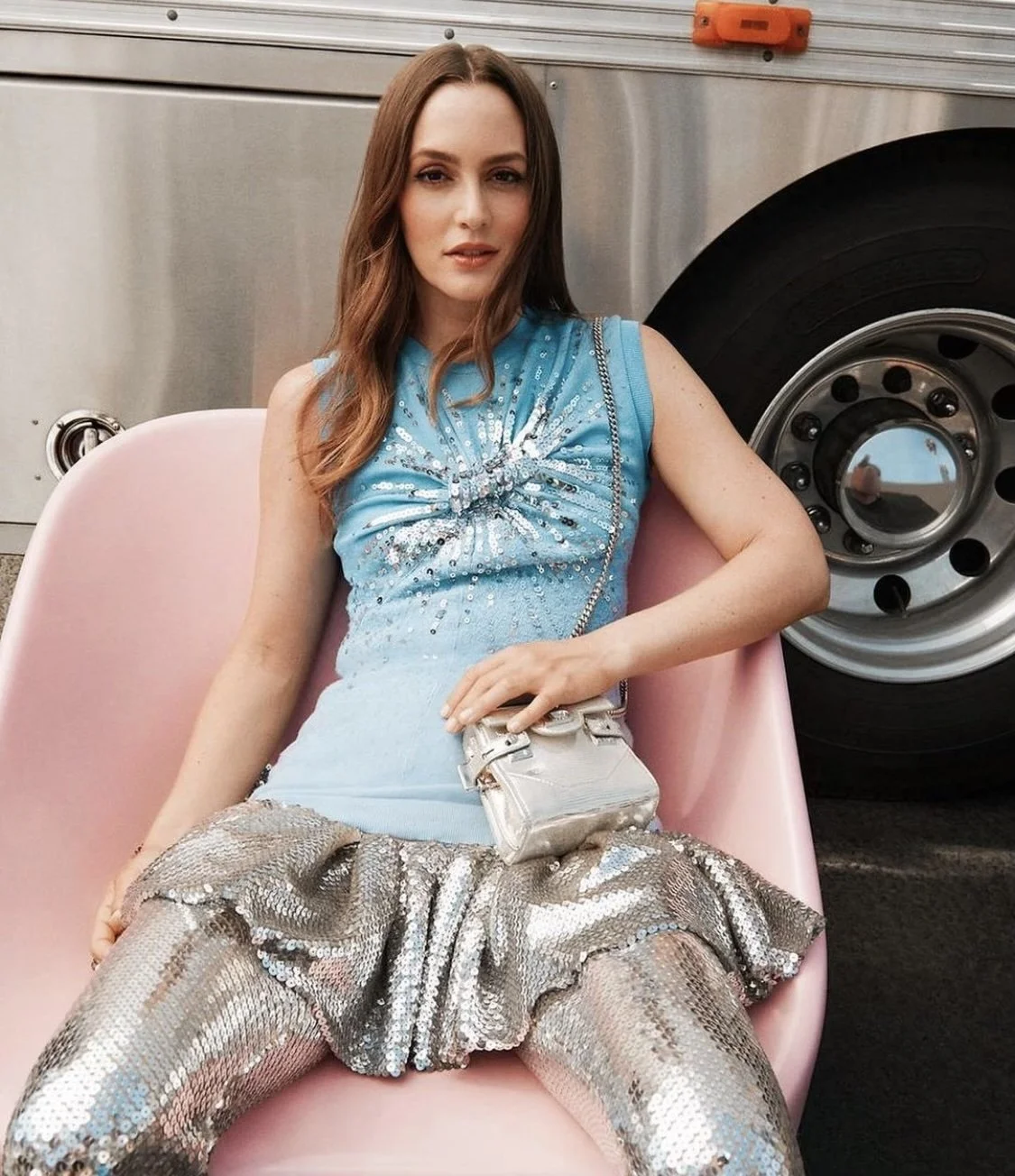 A fair-skinned woman with brown hair, wearing a blue, sequined top and shiny pants, poses on a pink chair in front of a silver bus.