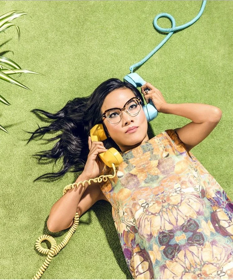 Woman lies on green surface and holds two retro telephones, one to each ear, wearing a patterned vintage dress and eyeglasses.