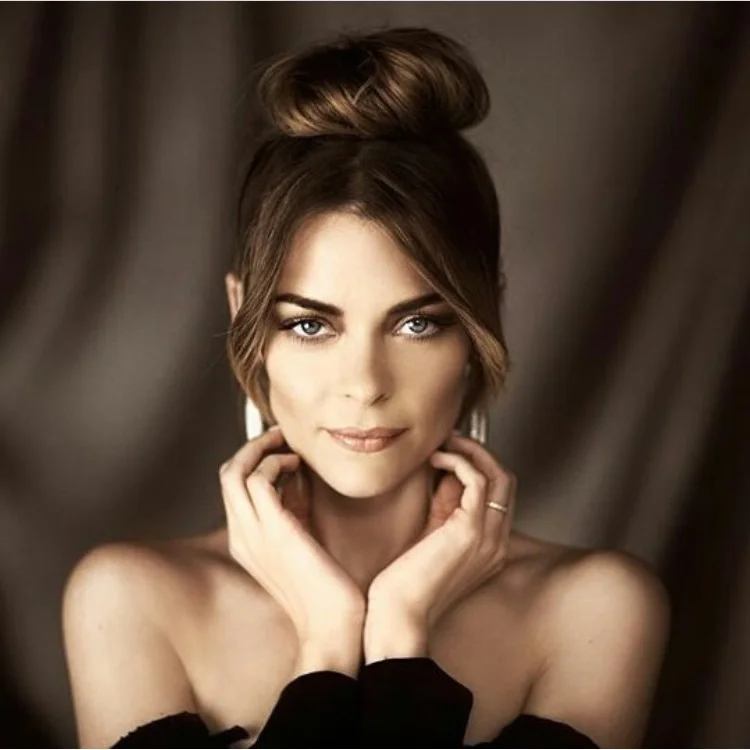 Close-up portrait of a beautiful adult woman with blue eyes and a bun hairstyle, set against a blurred grey background.