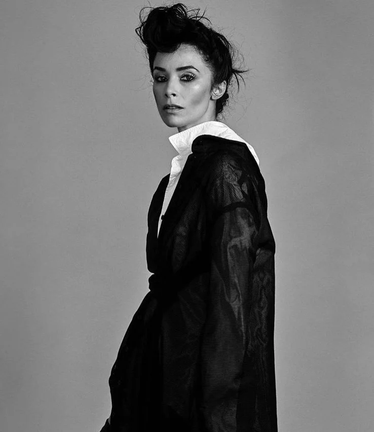 Black and white portrait of a woman with dramatic hair and makeup, wearing a sheer black robe over a white collared shirt against a neutral background.
