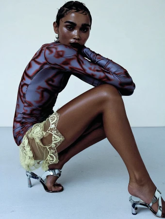 A model with wet hair poses in a long-sleeved top and yellow lace skirt, wearing silver high heels against a simple white background.