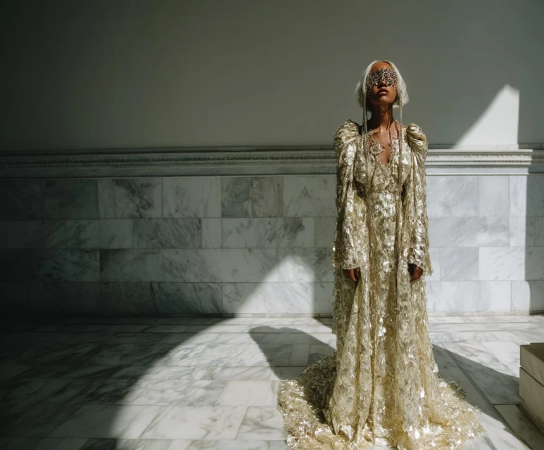 Woman in long, embellished gold dress and embellished mask poses in a brightly lit marble-lined interior. Shadows create contrast.