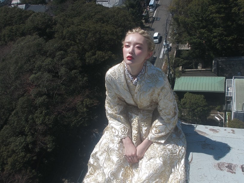Fashionable young woman in gold gown poses on rooftop, surrounded by dense trees and a distant urban street on a sunny day.