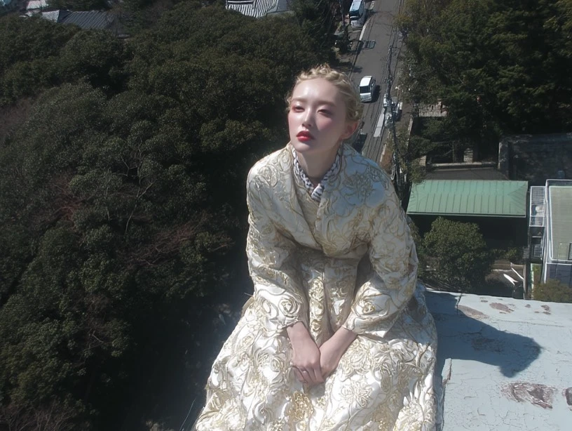 Fashionable young woman in gold gown poses on rooftop, surrounded by dense trees and a distant urban street on a sunny day.