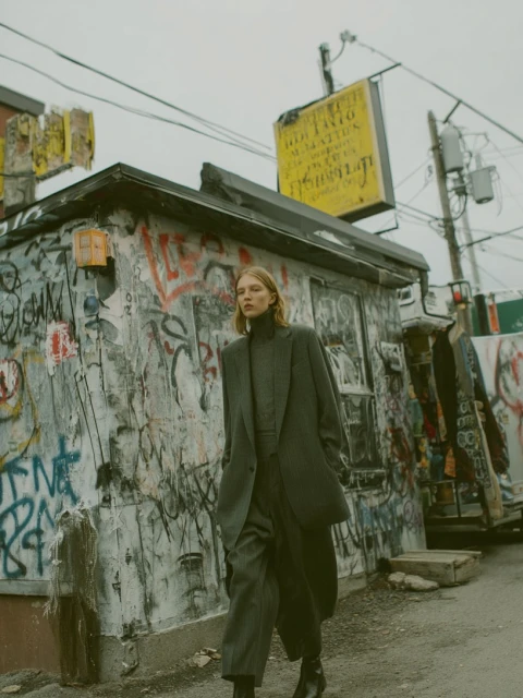 Woman in an overcoat and matching suit walks on an urban street in front of a graffitied building under an overcast sky.