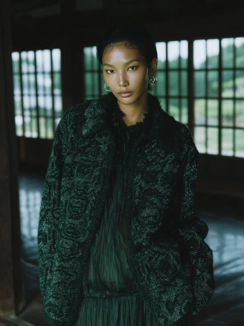 Portrait of a young woman wearing a dark green textured jacket and pleated dress, posing indoors with natural light.