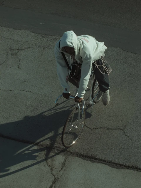 Overhead shot of a person riding a silver bicycle on cracked asphalt. The rider wears a hoodie and dark pants with chains.
