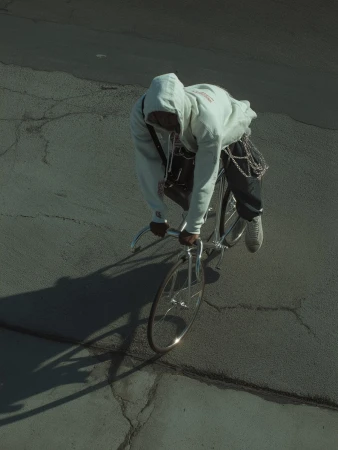 Overhead shot of a person riding a silver bicycle on cracked asphalt. The rider wears a hoodie and dark pants with chains.