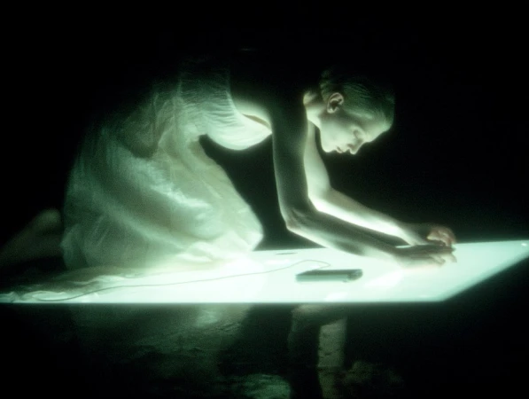Woman in a light-colored dress kneels and leans over a glowing white surface in a dark room, with a dark object on the surface.