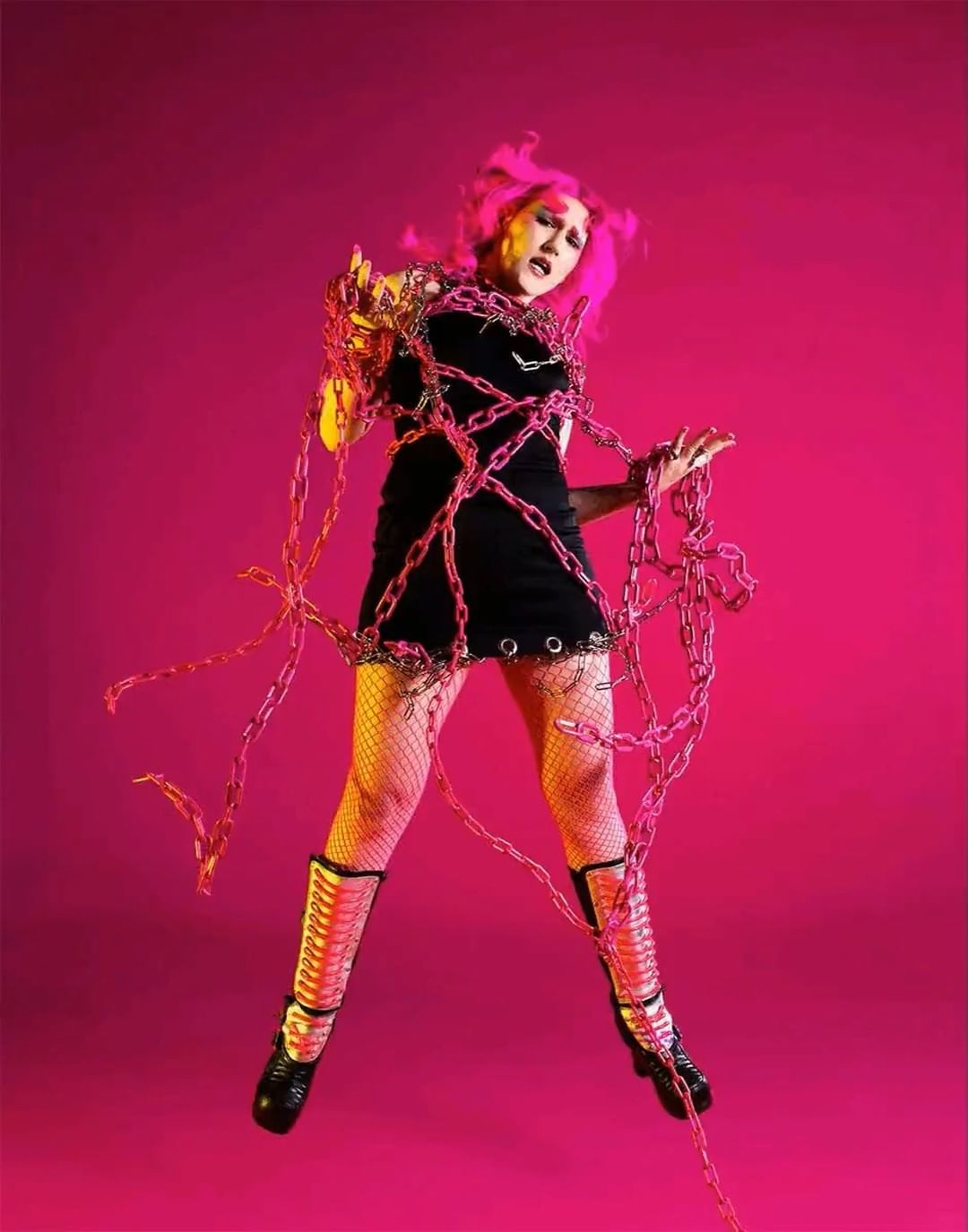 A woman with pink hair poses indoors in a black dress and boots, covered with metal chains, jumping against a pink backdrop.