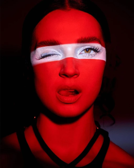 Close-up studio shot of a winking woman with painted red and white stripes across her face, with a black, strappy top visible.