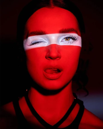 Close-up studio shot of a winking woman with painted red and white stripes across her face, with a black, strappy top visible.