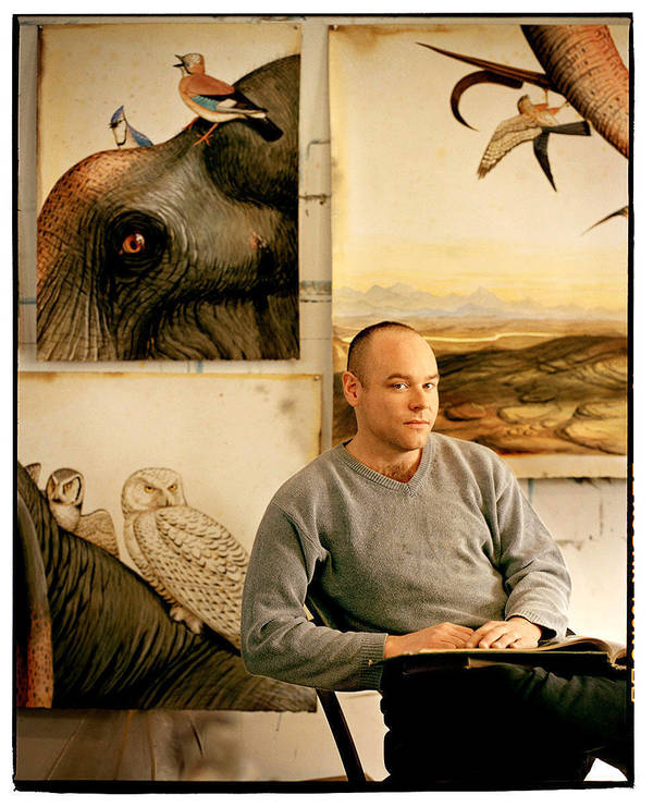 Adult male artist poses in photo studio with painted canvases showing various birds and an elephant. Calm studio portrait.