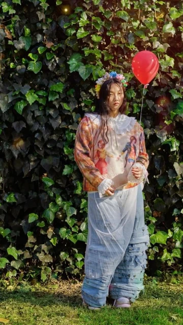 Young woman in decorative sweater and wide leg jeans stands in a garden with a red balloon in an outdoor setting.