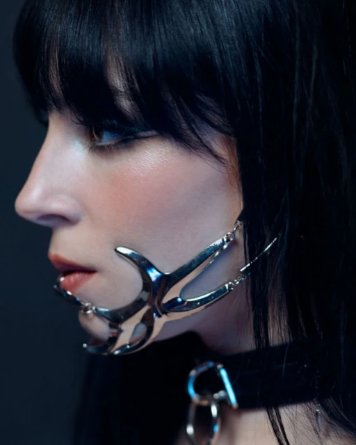 Adult female in a close-up studio portrait, wearing a decorative metal face piece and black choker, against a dark, gradient backdrop.